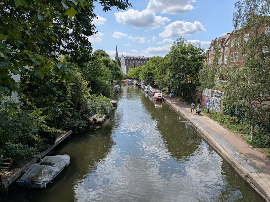 A scenic view of a narrow, calm canal in Paddington W2, lined with lush green trees on both sides. Along the right bank, a concrete pathway runs parallel to the water, with a few pedestrians strolling