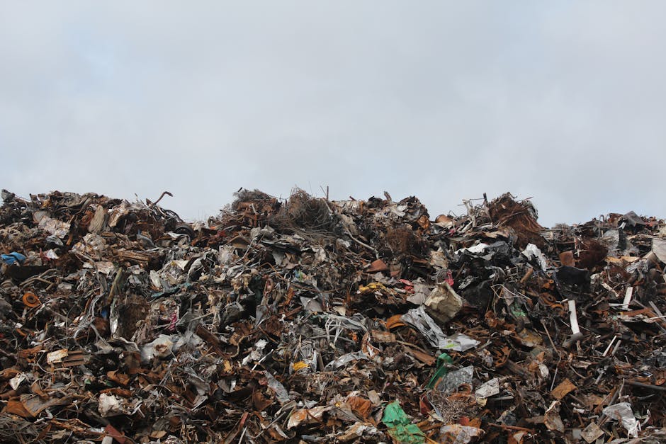 A large mound of assorted waste materials including discarded paper, plastic, metal scraps, and organic debris under a cloudy sky, illustrating the environmental impact of improper waste disposal dist