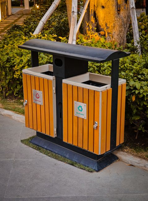 A dual-compartment outdoor waste and recycling bin situated on a paved sidewalk, featuring orange wooden panel fronts with silver handles and labels indicating 'trash' and 'recycling.' The bin has a b