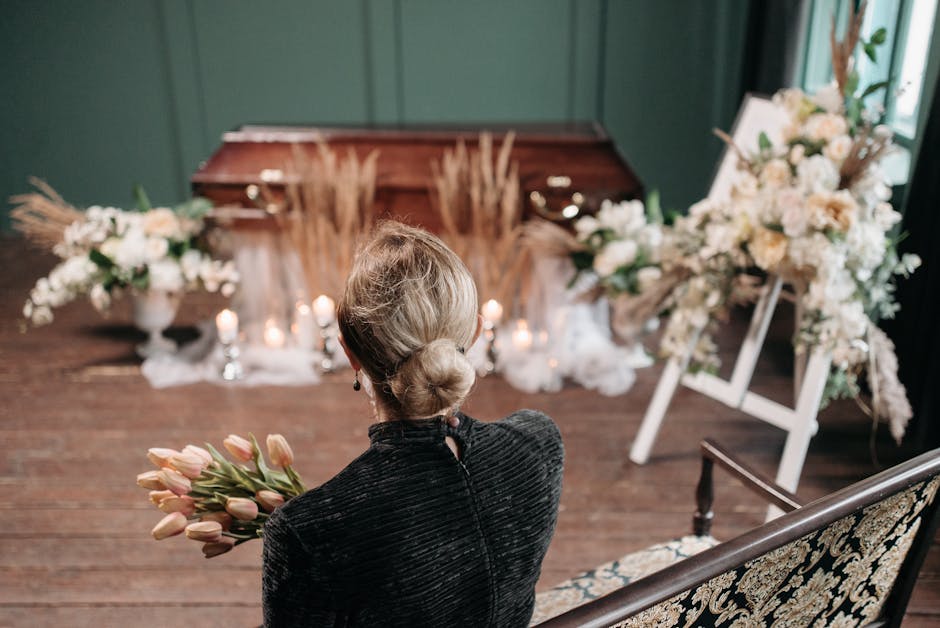 A woman with her hair styled in a low bun, wearing a dark textured jacket, is holding a bouquet of pink tulips in front of her. She faces away from the camera, seated on an ornate, upholstered bench. 