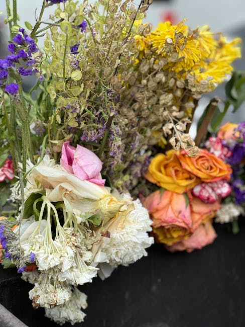 An arrangement of fresh flowers including white, cream, and pink roses, complemented by yellow, purple, and lavender blossoms, with some flowers slightly wilted. The bouquet features a mixture of text