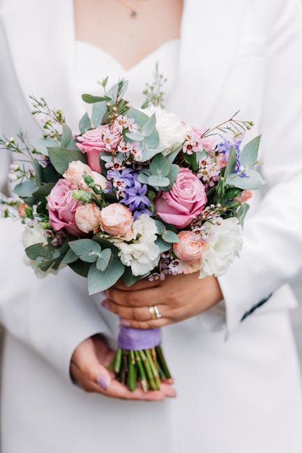 A person dressed in a white blazer is holding a bridal bouquet composed of pink roses, peach-colored spray roses, white lisianthus, and sprigs of blue-purple delphiniums, accented with green eucalyptu