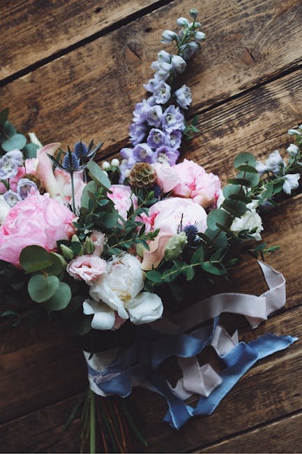 A floral bouquet resting on a rustic wooden surface, featuring light pink peonies, white ranunculus, soft blue delphiniums, and sprigs of greenery including eucalyptus. The arrangement is wrapped with