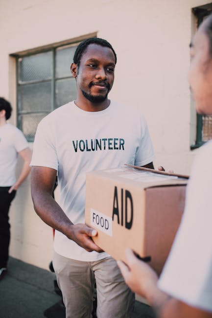 A volunteer with dark skin, facial hair, and wearing a white T-shirt with 'VOLUNTEER' printed on it, is seen handing a cardboard box labeled 'AID' and 'FOOD' to a woman whose face is partially visible