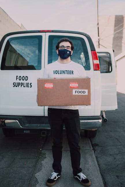 A man wearing glasses and a navy blue face mask stands outdoors in front of a white van marked 'FOOD SUPPLIES' and 'FOOD,' holding a large cardboard box labeled 'FOOD' with red and white stickers. The